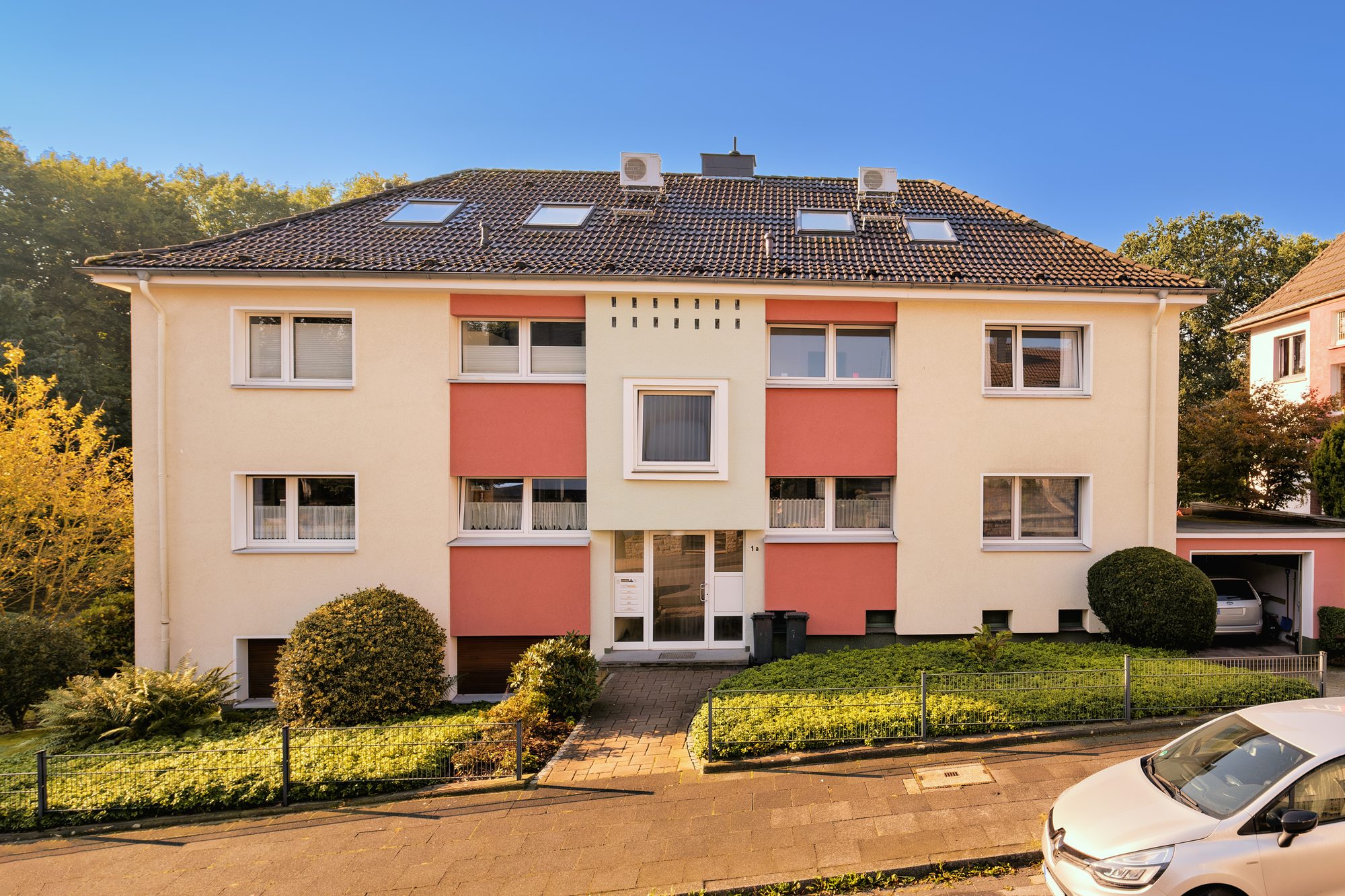 Mehrfamilienhaus mit roten Putzfeldern und Walmdach hinter einer Hecke